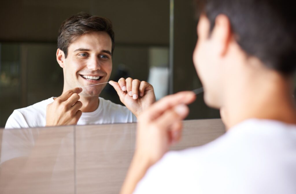 Smiling man with brown hair and white T-shirt flossing in front of a mirror.