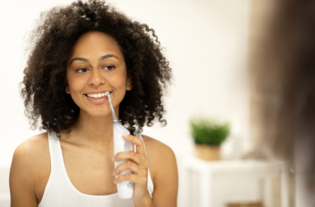 Woman with dark curly hair in a white tank top using water flosser on teeth.