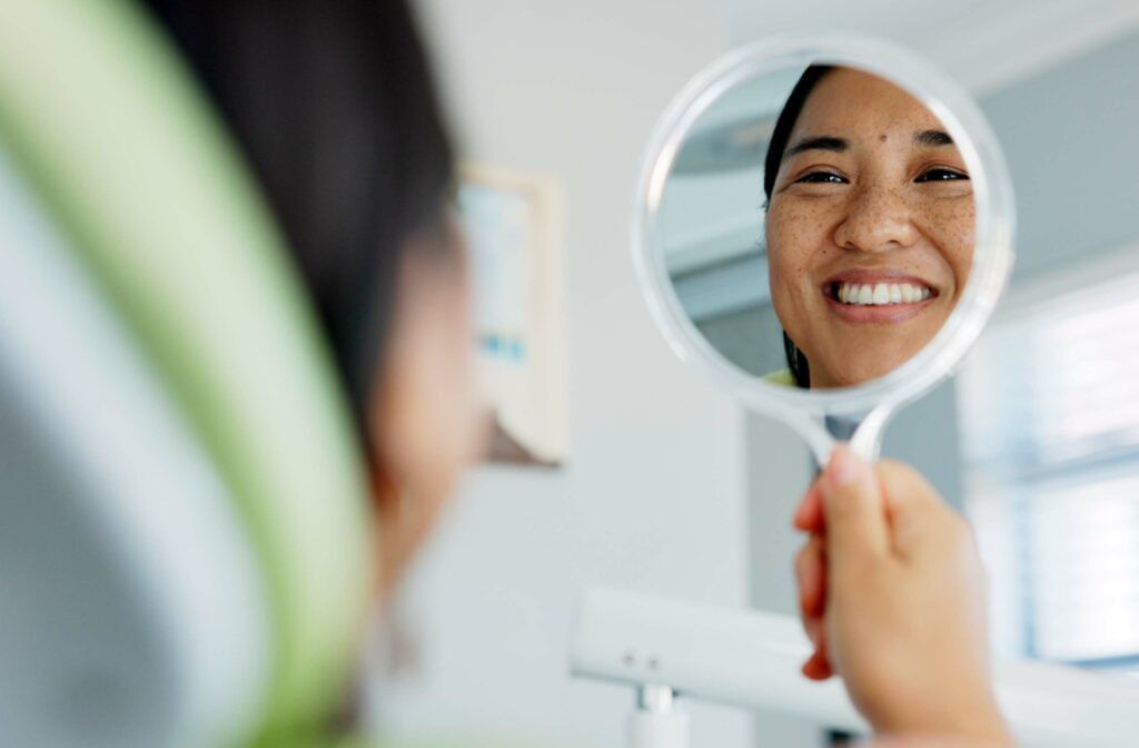 Woman smiling and looking at her teeth in a handheld mirror after a dental treatment.
